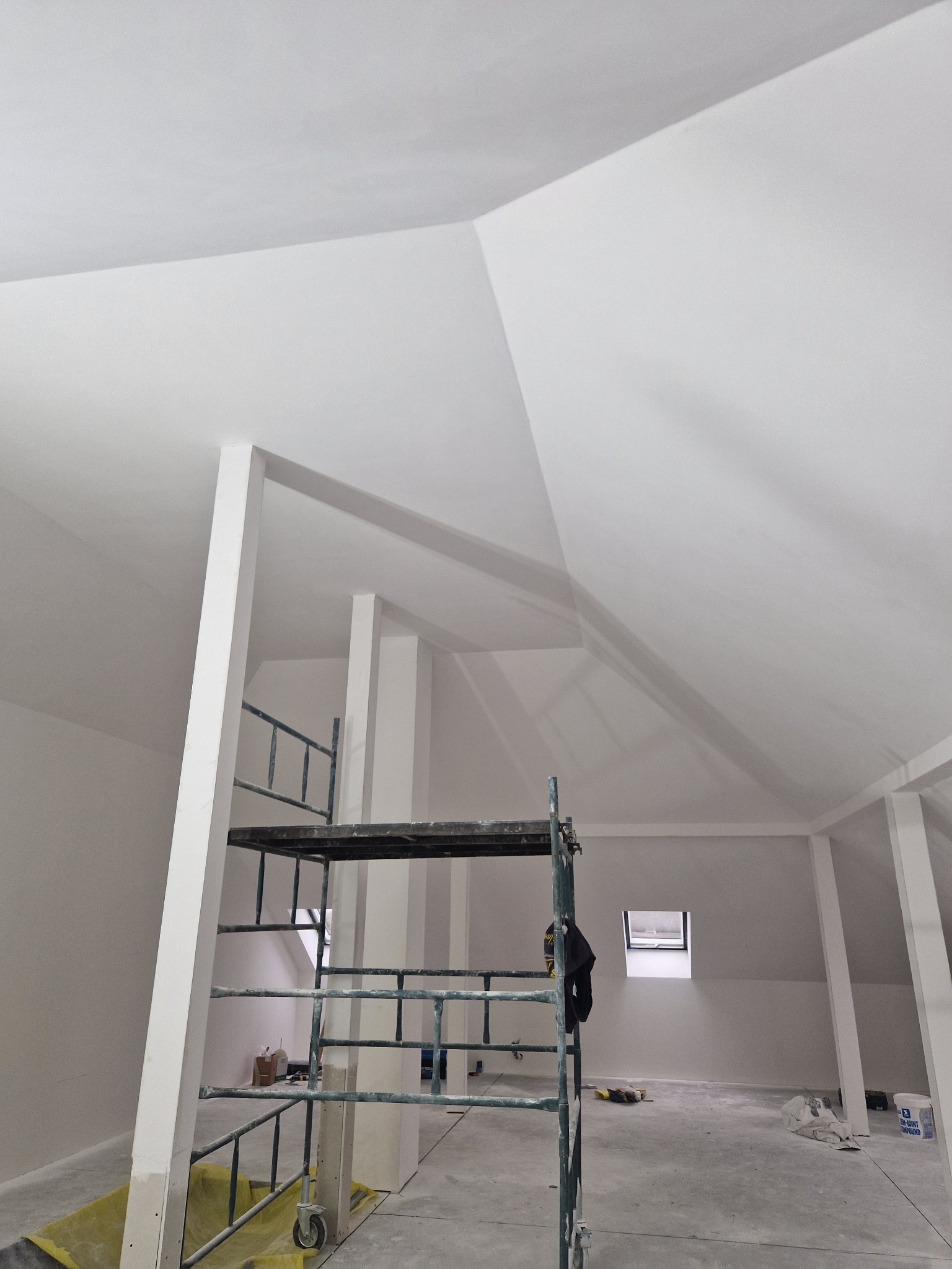 Poddasze po pracach wykończeniowych, ściany i sufit pomalowane na biało, widoczne rusztowanie, małe okno dachowe, kolumny podtrzymujące strop.