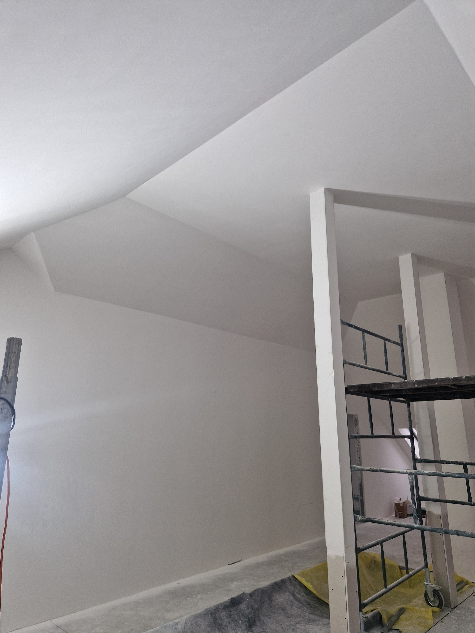 Pokój w trakcie wykańczania, świeżo pomalowane ściany i sufit w odcieniach bieli, widoczne rusztowanie i zabezpieczenia podłogi, skos sufitu.