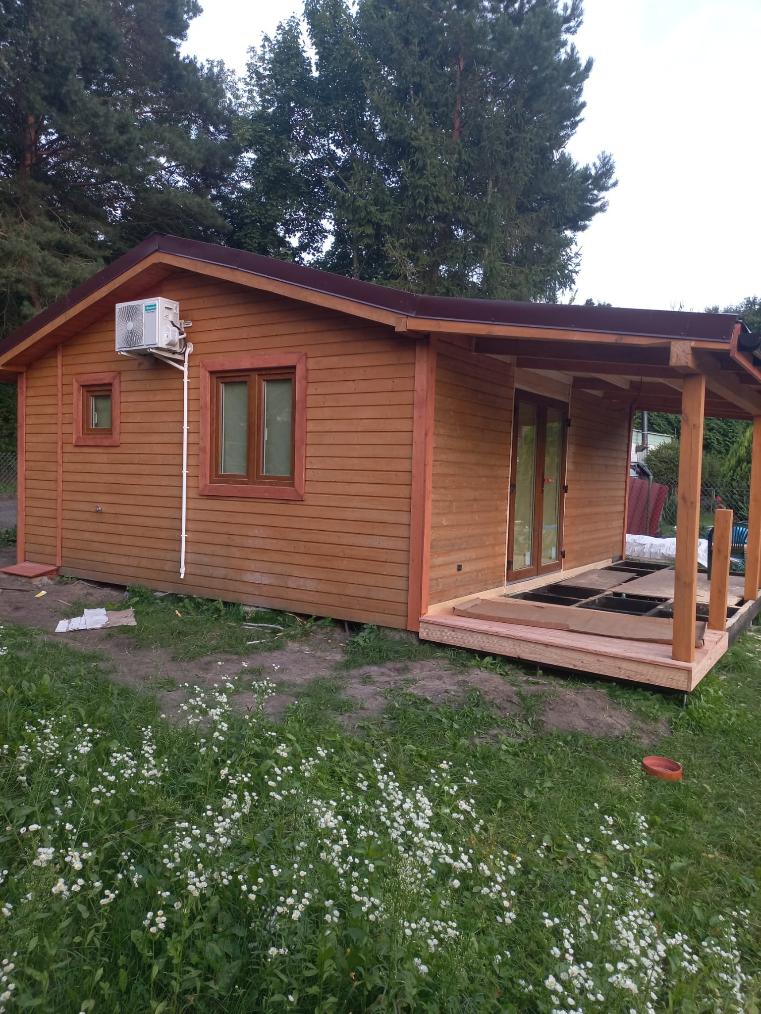 Brązowy domek szkieletowy z klimatyzatorem na elewacji, w trakcie budowy werandy. Widoczne elementy konstrukcyjne i podłoże pod taras.