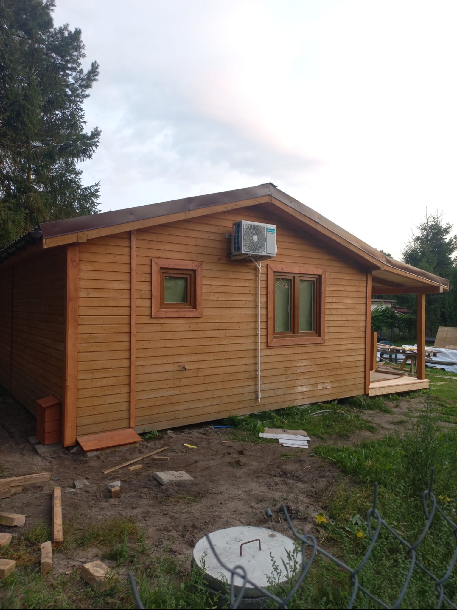 Z zewnątrz: drewniany domek z brązowym dachem i klimatyzacją na ścianie. Widoczne okna z brązowymi ramami i fragment tarasu. Teren wokół domu częściowo zagospodarowany.