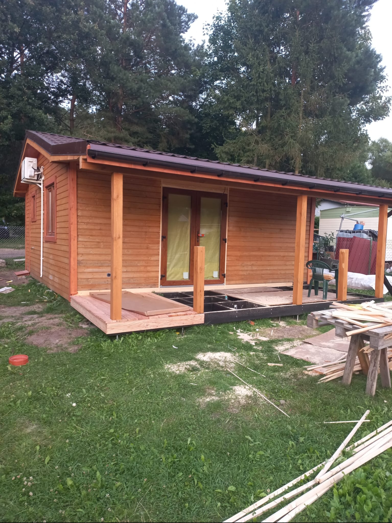 Drewniany domek letniskowy w trakcie budowy, z widoczną konstrukcją tarasu i elementami wykończeniowymi. W tle drzewa i zarys innego budynku.