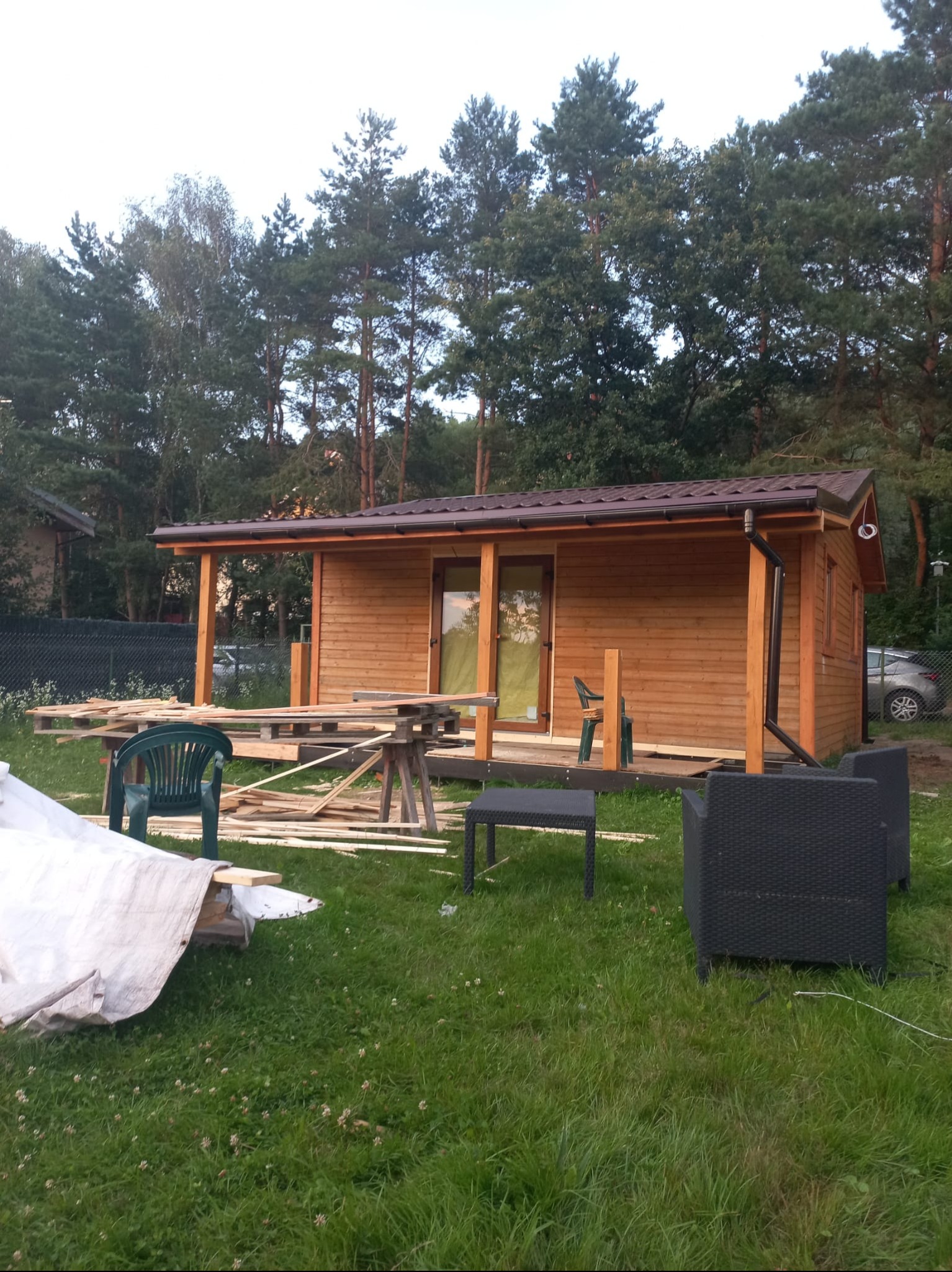 Niedokończony drewniany domek letniskowy z brązowym dachem na zewnątrz, otoczony zielenią i meblami ogrodowymi. Część materiałów budowlanych leży obok domku.