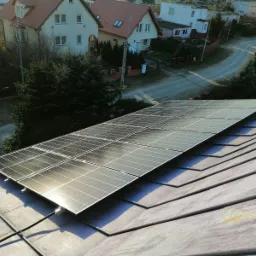 Panele słoneczne zamontowane na szarym, spadzistym dachu domu jednorodzinnego w słoneczny dzień, widok z g&oacute;ry na okoliczną zabudowę.