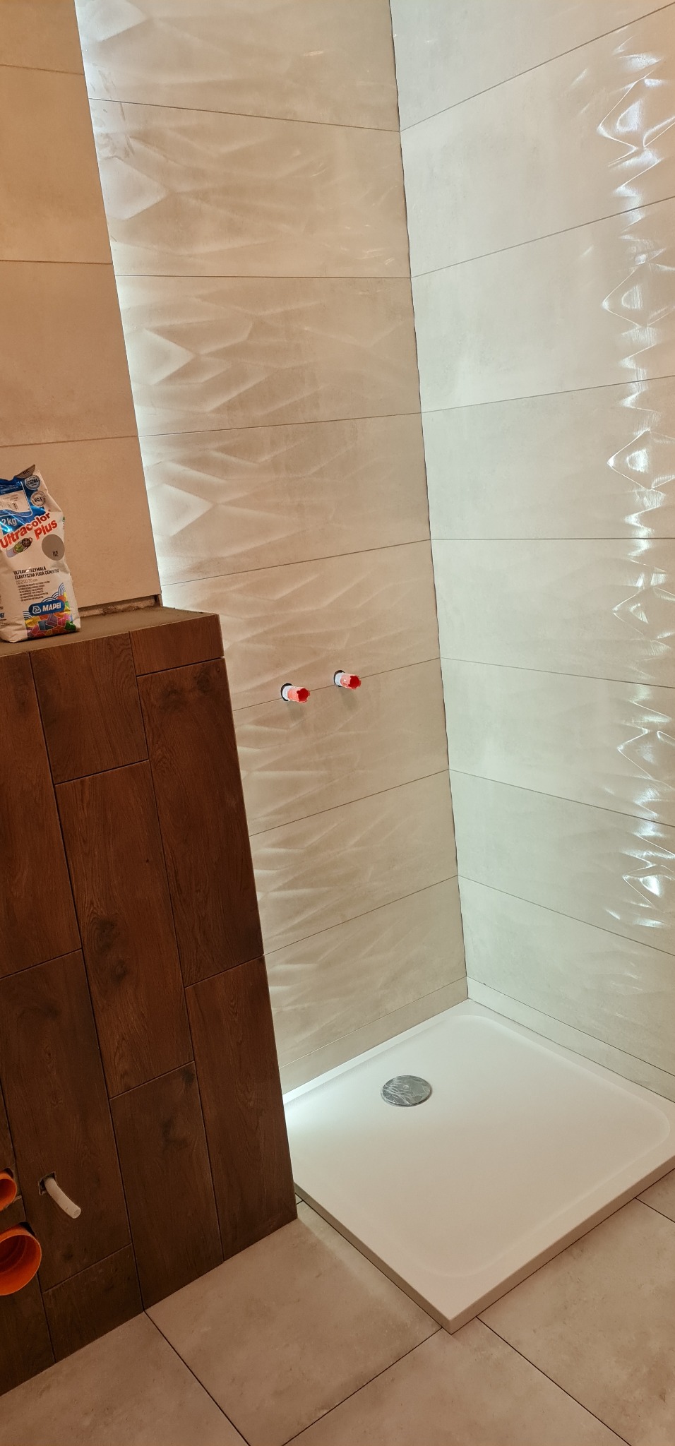 Nowoczesna kabina prysznicowa z brodzikiem i odpływem liniowym, wykończona beżowymi płytkami z delikatnym wzorem, widoczny worek z zaprawą do fugowania i niezabudowane przyłącza wodne.