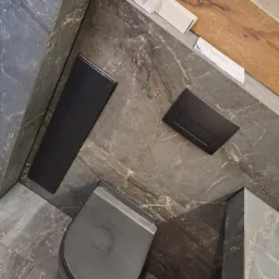 Zawieszona miska WC w odcieniach szarości, z czarnym panelem spłukującym i czarnym grzejnikiem łazienkowym na tle marmurowych płytek z złotymi żyłkami. Na górze widoczne pudełka po lampach...