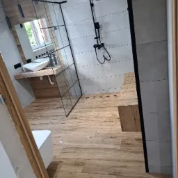Nowoczesna łazienka z drewnopodobną podłogą, szarą glazurą, kabiną prysznicową typu walk-in z czarnymi profilami, umywalką nablatową na drewnianym blacie i czarną armaturą.