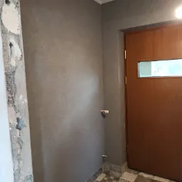 Szara ściana o strukturze baranka po położeniu gładzi, widoczne fragmenty starej ściany przed obróbką, dekoracyjna podłoga z płytek w stylu patchwork, brązowe drzwi z małym okienkiem.