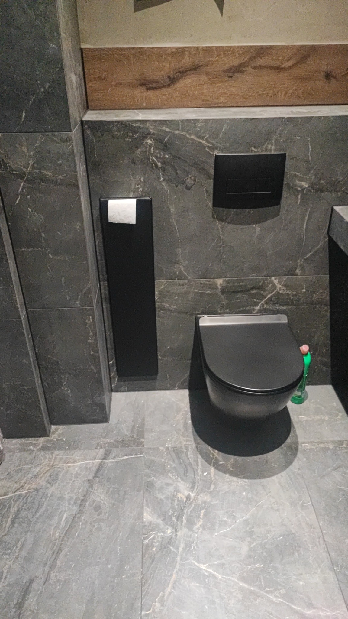 Nowoczesna toaleta podwieszana w łazience wykończonej płytkami imitującymi marmur, z czarnym pojemnikiem na papier toaletowy i przyciskiem spłukującym w tym samym kolorze. Na podłodze szare płytki...