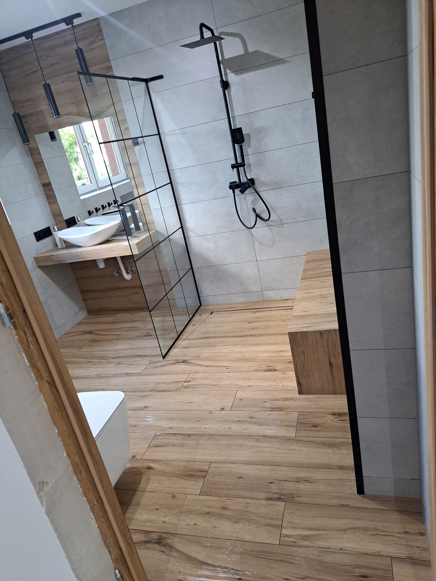 Nowoczesna łazienka z drewnopodobną podłogą, szarą glazurą, kabiną prysznicową typu walk-in z czarnymi profilami, umywalką nablatową na drewnianym blacie i czarną armaturą.