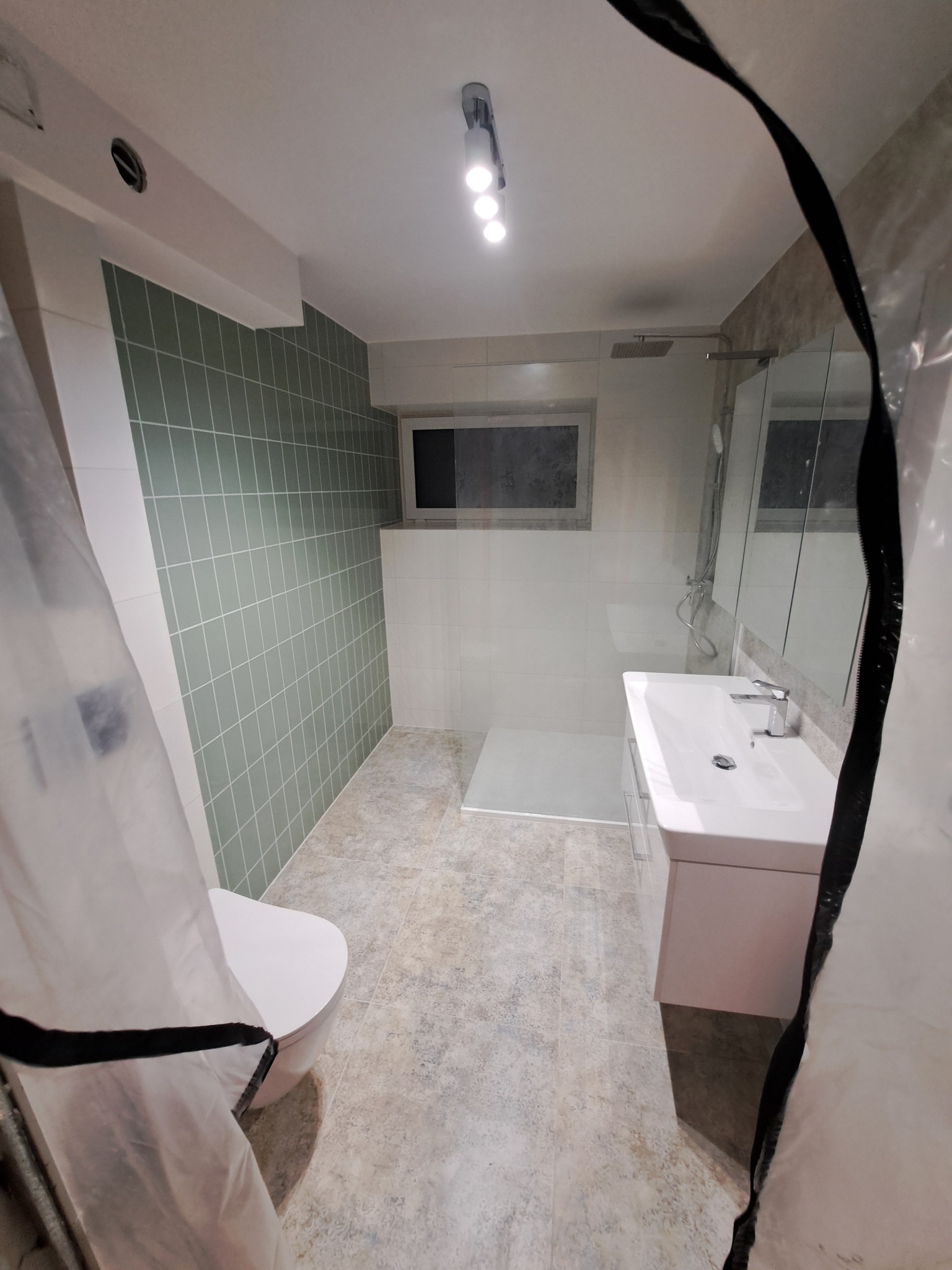 Ukończona łazienka z zielonymi i białymi płytkami, kabiną prysznicową, umywalką z szafką i toaletą, częściowo zasłonięta folią ochronną.