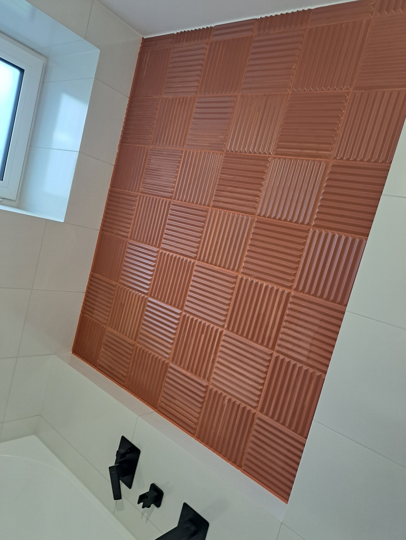 Ściana łazienki wyłożona dekoracyjnymi, brązowymi płytkami 3D w kwadraty, z widoczną białą armaturą i oknem.