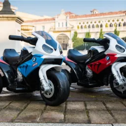 Dwa motocykle elektryczne BMW S1000RR dla dzieci, jeden niebieski, drugi czerwony, stojące na brukowanym dziedzińcu z budynkiem w tle.