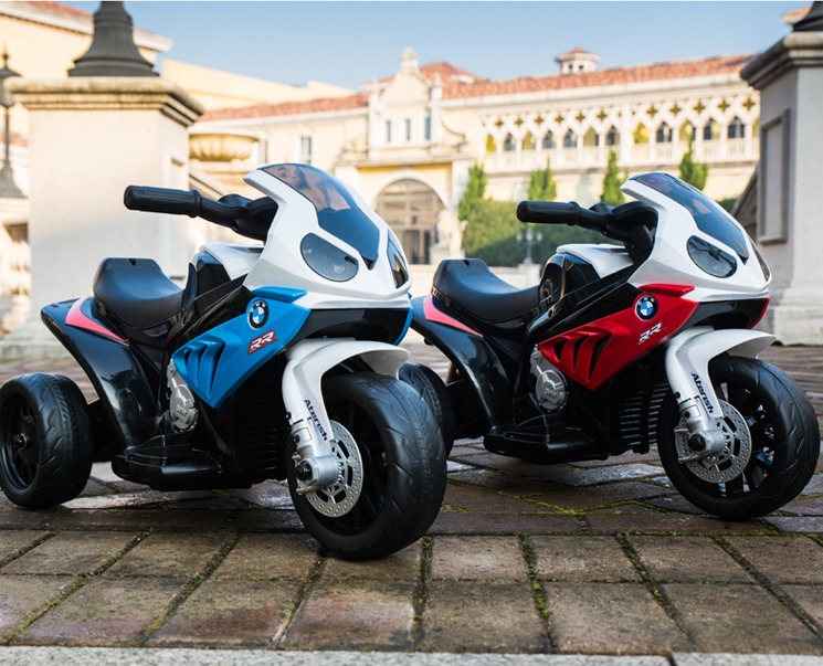 Dwa motocykle elektryczne BMW S1000RR dla dzieci, jeden niebieski, drugi czerwony, stojące na brukowanym dziedzińcu z budynkiem w tle.