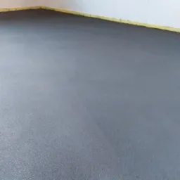 Świeżo wylana, gładka, szara posadzka jastrychowa, zabezpieczona żółtą taśmą przy ścianach, widoczne dwa gniazdka elektryczne w górnym rogu ściany.