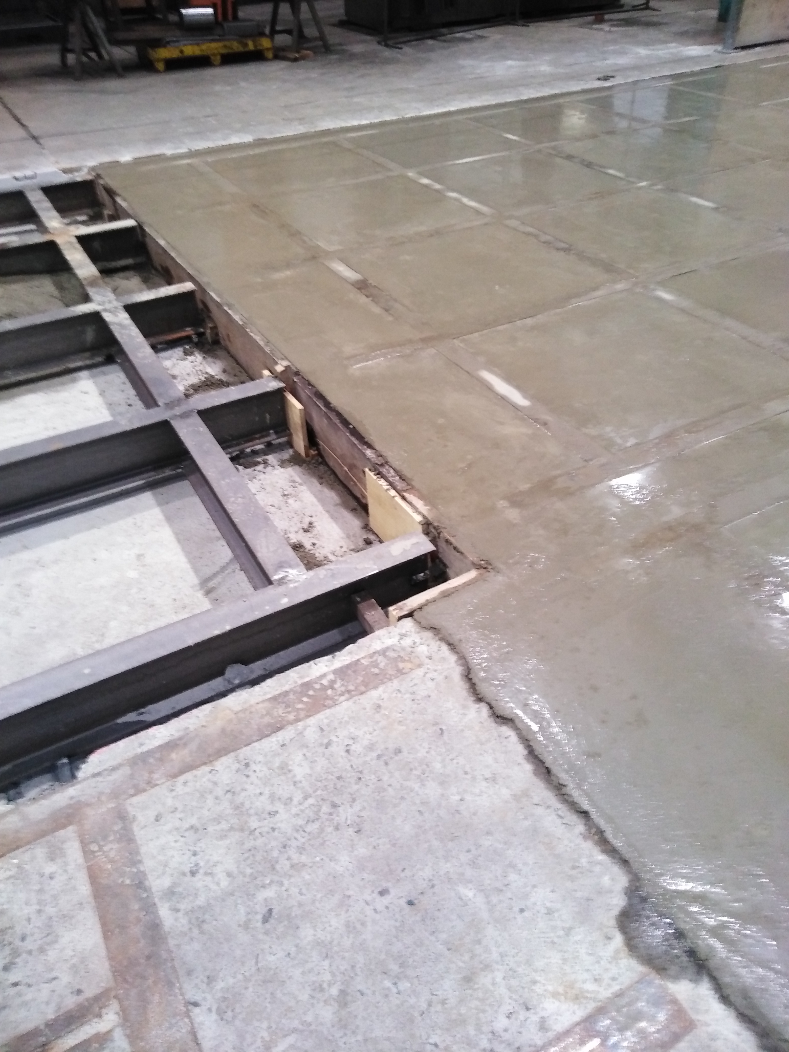 Świeżo wylany jastrych na podłodze przemysłowej z widocznymi metalowymi profilami i drewnianym szalunkiem, tworzący kwadratowe sekcje.