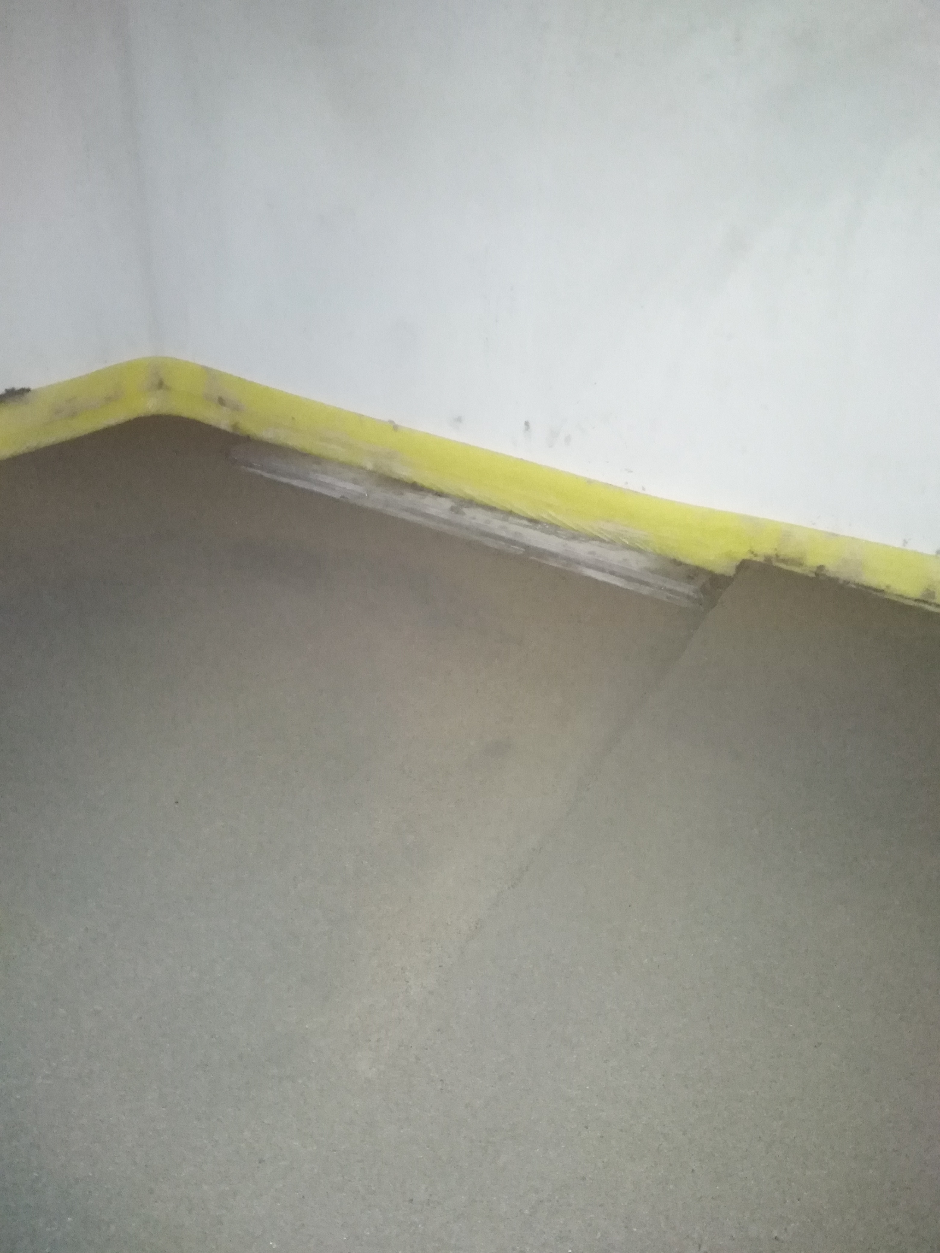 Świeżo wylana, szara posadzka z widocznym żółtym zabezpieczeniem przy ścianie, gotowa do dalszych prac wykończeniowych.