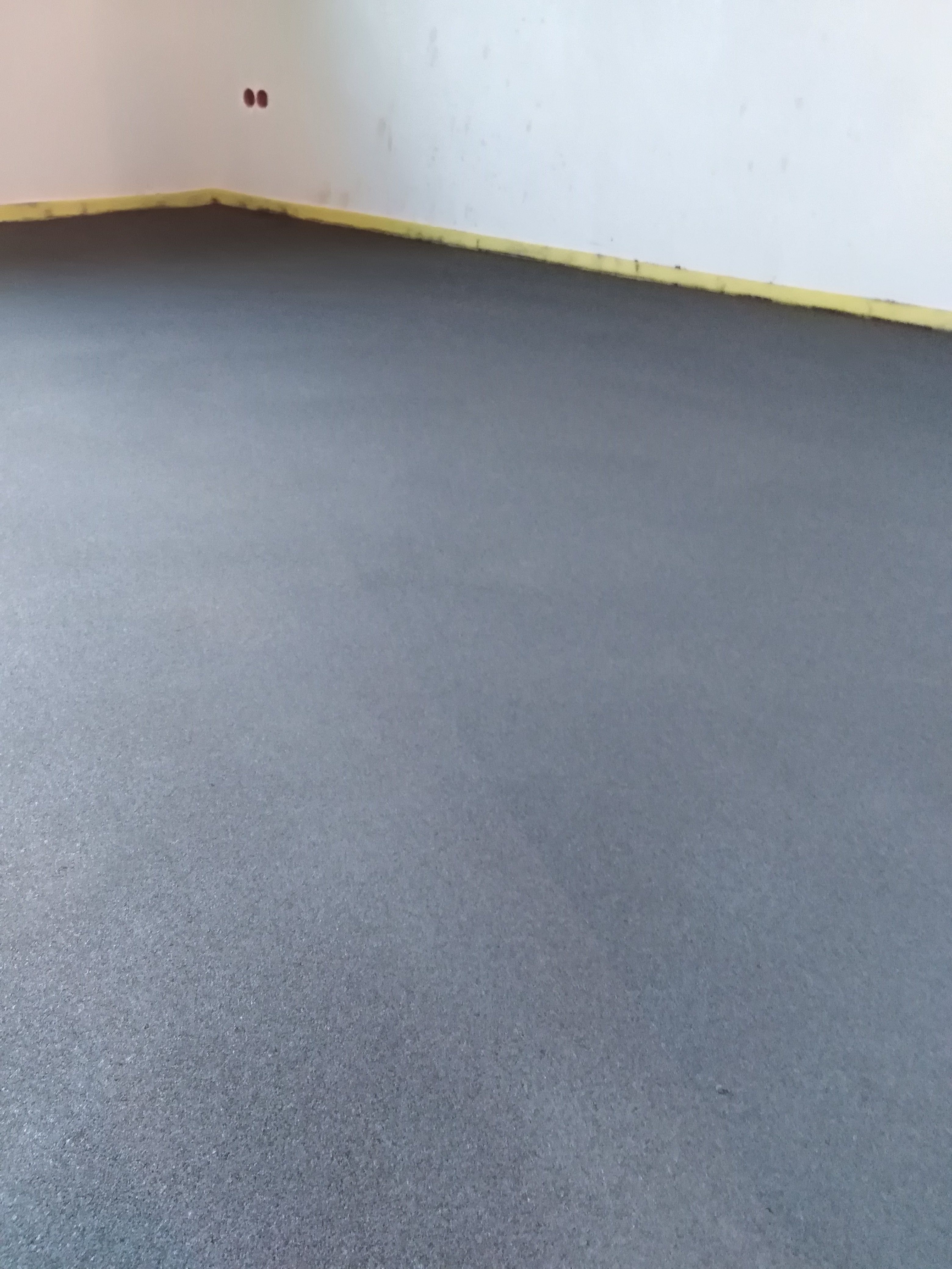 Świeżo wylana, gładka, szara posadzka jastrychowa, zabezpieczona żółtą taśmą przy ścianach, widoczne dwa gniazdka elektryczne w górnym rogu ściany.