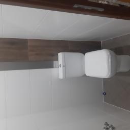 Luk-Bud - Biała toaleta w wykończonej łazience z białymi i brązowymi płytkami, widoczne fragmenty instalacji wodnej.