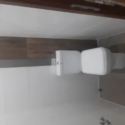 Biała toaleta w wykończonej łazience z białymi i brązowymi płytkami, widoczne fragmenty instalacji wodnej.