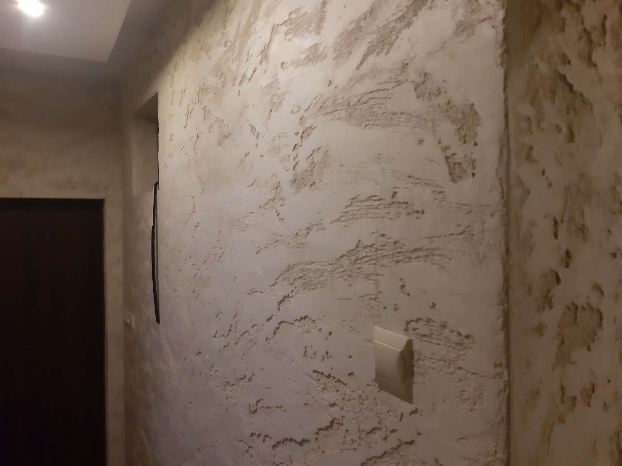 Dekoracyjna ściana o fakturze trawertynu w korytarzu, widoczny włącznik światła i fragment drzwi.