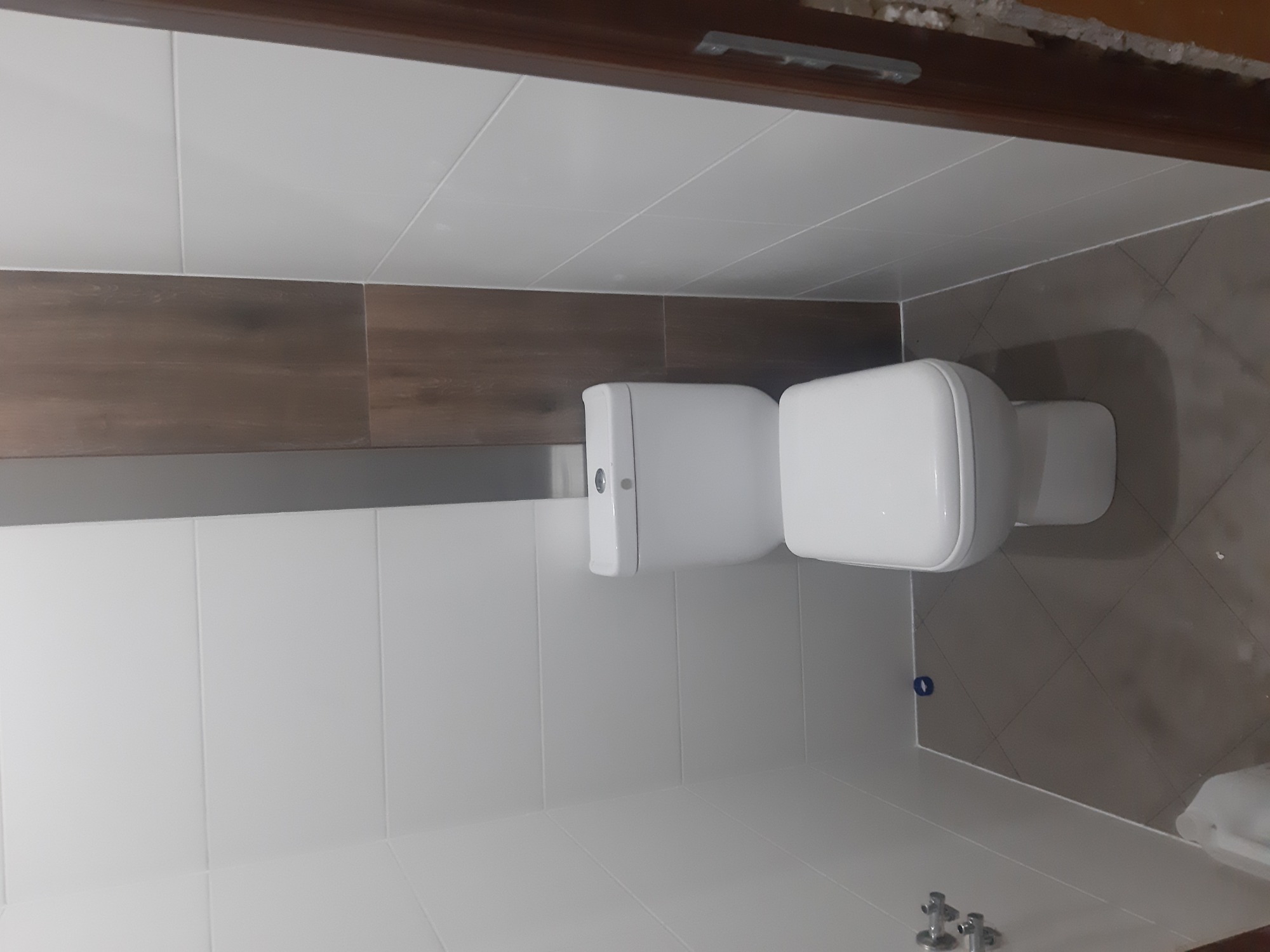 Biała toaleta w wykończonej łazience z białymi i brązowymi płytkami, widoczne fragmenty instalacji wodnej.