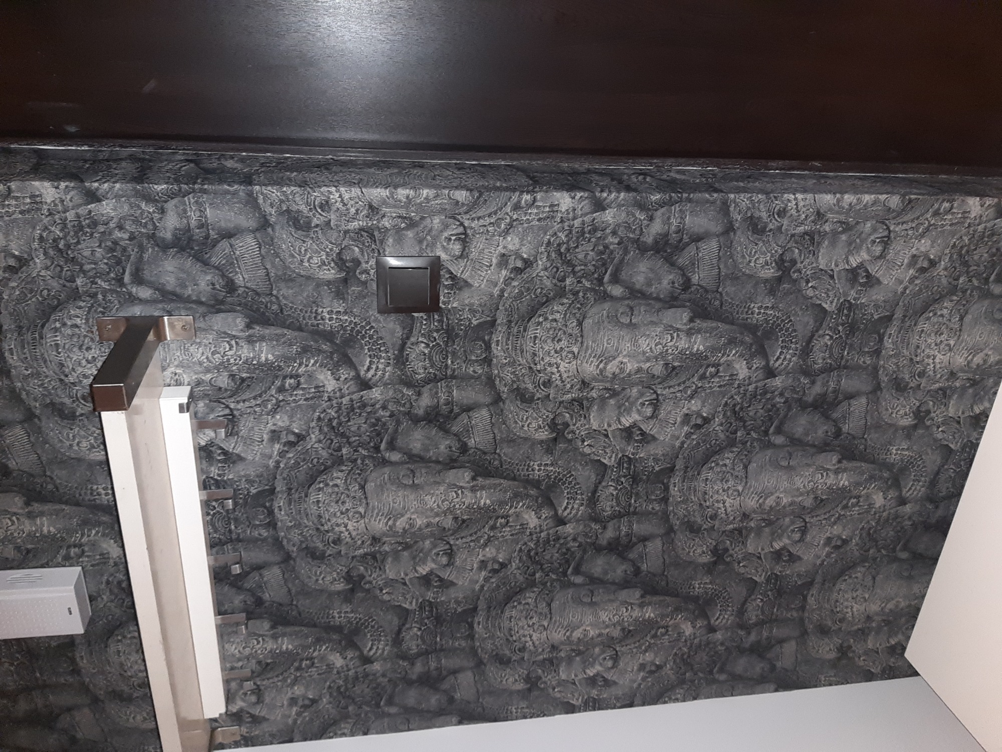 Ściana pokryta tapetą w odcieniach szarości z motywem reliefów, widoczny włącznik światła i fragment karnisza.