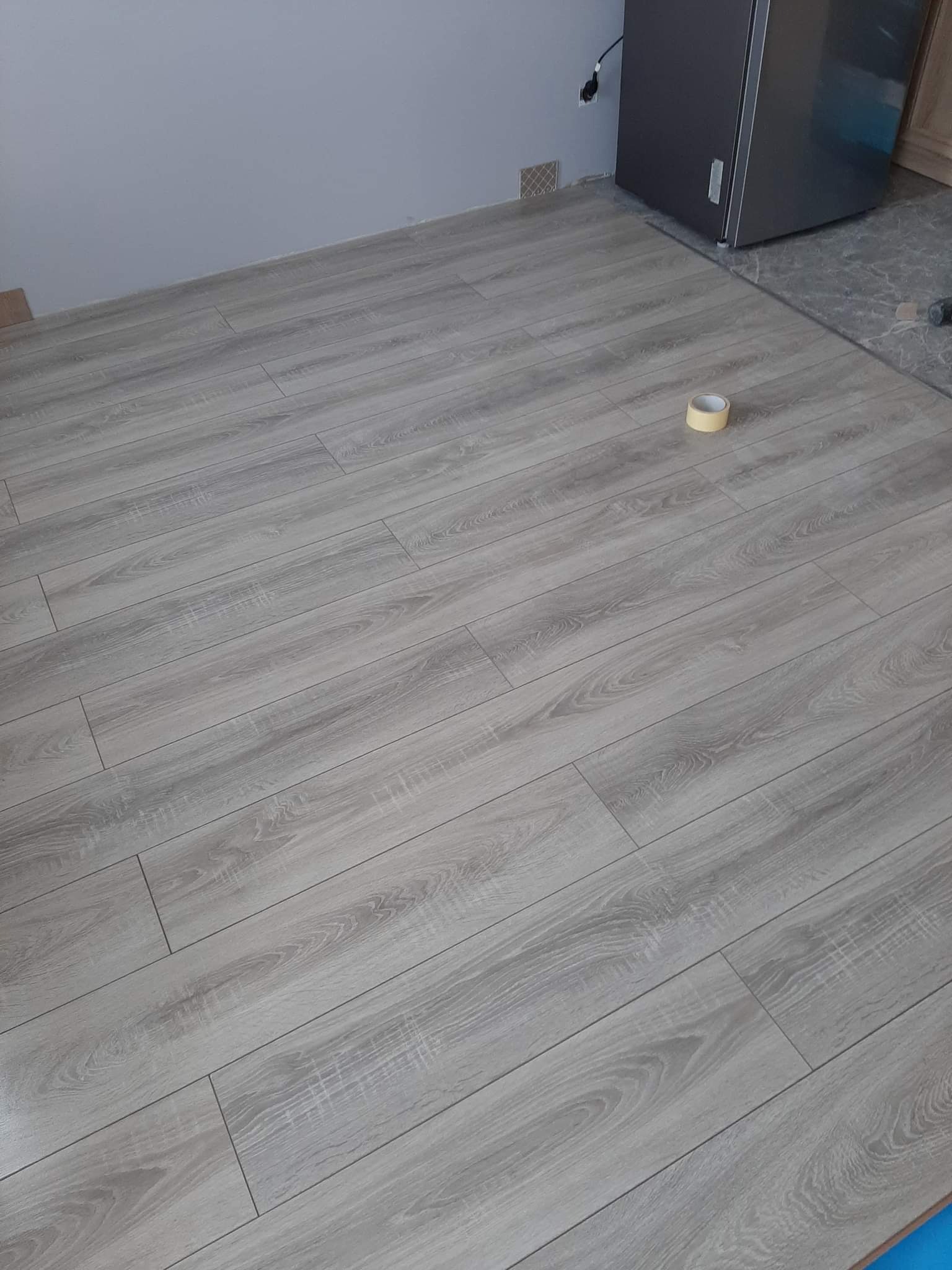 Ułożone panele podłogowe w odcieniach szarości, widoczna rolka taśmy malarskiej, fragment ściany i lodówki