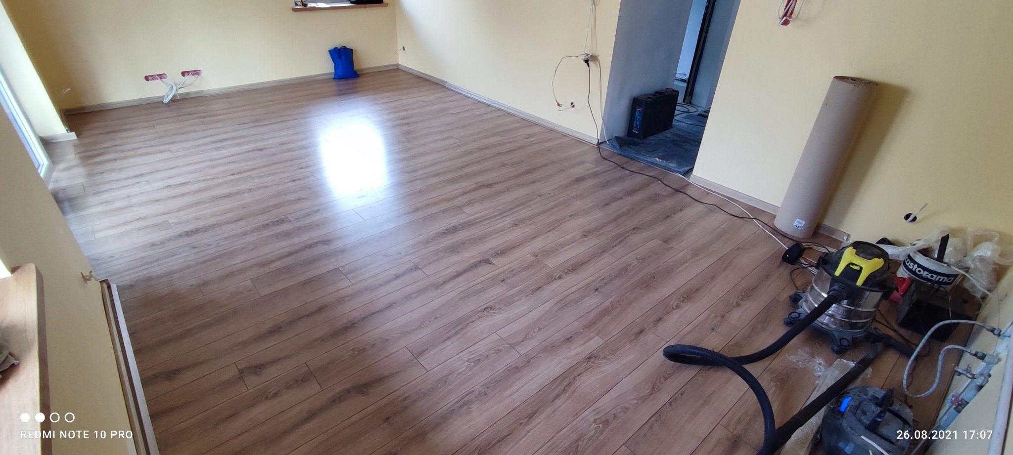 Nowo położona podłoga z paneli drewnopodobnych w jasnym pokoju, widoczny odkurzacz przemysłowy i resztki materiałów instalacyjnych przy ścianie.