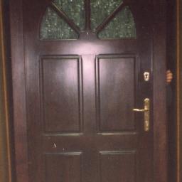 ADAM ARS MEBLE LITE DREWNO RENOWACJE. - Ciemnobrązowe drzwi wejściowe z półokrągłym przeszkleniem w górnej części, widoczne zamek i klamka, fragment dłoni po prawej stronie.