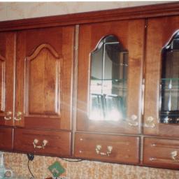 ADAM ARS MEBLE LITE DREWNO RENOWACJE. - Wisząca szafka kuchenna z ciemnego drewna z przeszklonymi drzwiczkami i ozdobnymi uchwytami, widoczne naczynia wewnątrz, na tle płytek ceramicznych.