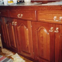 ADAM ARS MEBLE LITE DREWNO RENOWACJE. - Dolna część zabudowy kuchennej z ciemnego drewna z ozdobnymi, złotymi uchwytami, widoczna torba na podłodze i nieporządek.