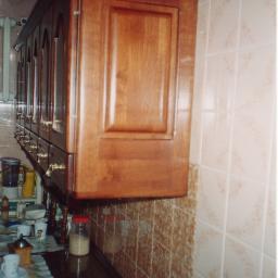 ADAM ARS MEBLE LITE DREWNO RENOWACJE. - Wisząca szafka kuchenna z drewna z frezowanymi frontami i złotymi uchwytami, zamontowana nad blatem kuchennym z widocznymi akcesoriami i wzorzystą ściereczką, tło stanowią płytki ceramiczne.