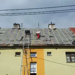 Pracownik w czerwonych spodniach na dachu budynku, montuje membranę dachową na drewnianej konstrukcji, widoczna drabina oparta o budynek, część dachu pokryta czerwoną dachówką.