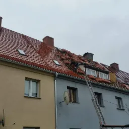 Pracownik na drabinie układa czerwoną dachówkę na dachu szeregowca w pochmurny dzień, widoczne okna dachowe.