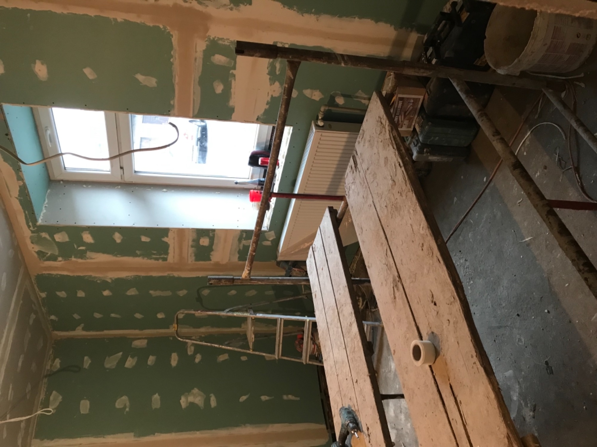 Wnętrze w trakcie remontu: ściany pokryte zieloną płytą gipsową, widoczne łączenia płyt, okno, grzejnik, rusztowanie i narzędzia na drewnianej desce.