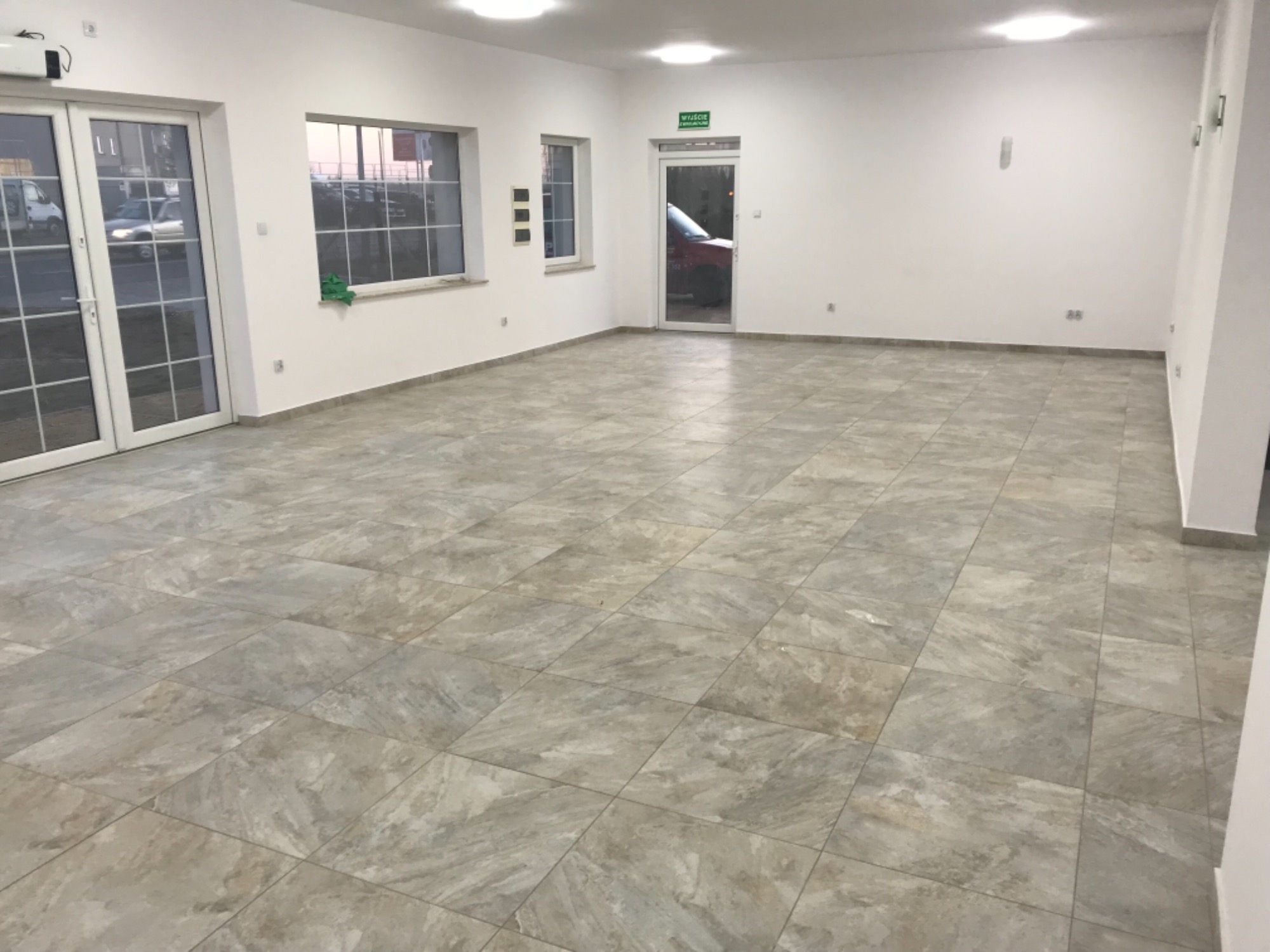 Wykończone wnętrze przestronnego pomieszczenia z nową, szarą podłogą z płytek imitujących kamień, białymi ścianami, dużymi oknami i drzwiami balkonowymi z widokiem na zewnątrz.