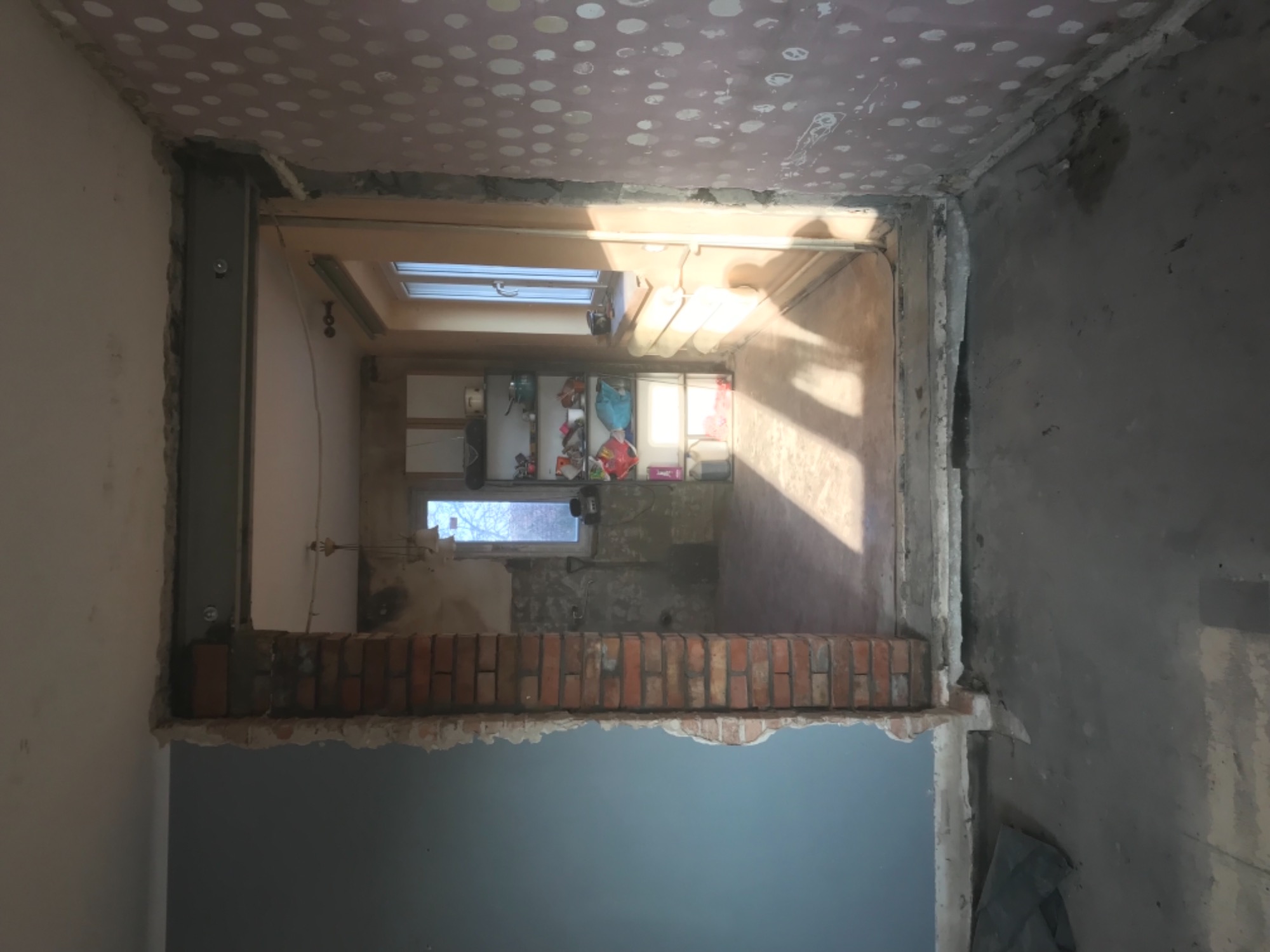 Wnętrze w trakcie przebudowy: widok z jednego pomieszczenia do drugiego przez otwór w ścianie, z widoczną cegłą, stalowym wzmocnieniem i częściowo wykończonym sufitem z widocznymi łatami. W oddali...