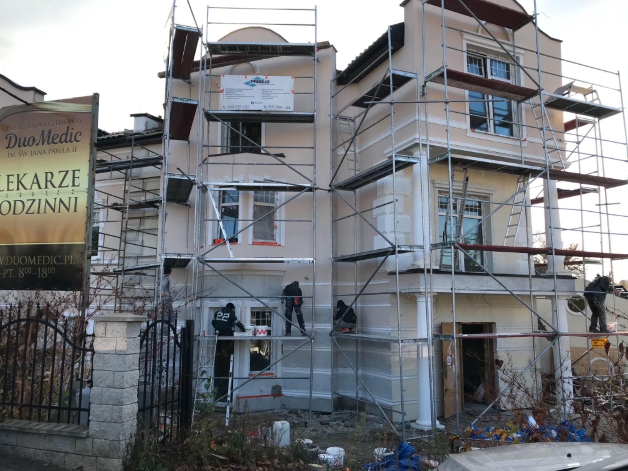 Dwupiętrowy budynek w trakcie prac elewacyjnych, otoczony rusztowaniami, z pracownikami w kombinezonach, widoczny szyld przychodni lekarskiej po lewej stronie.