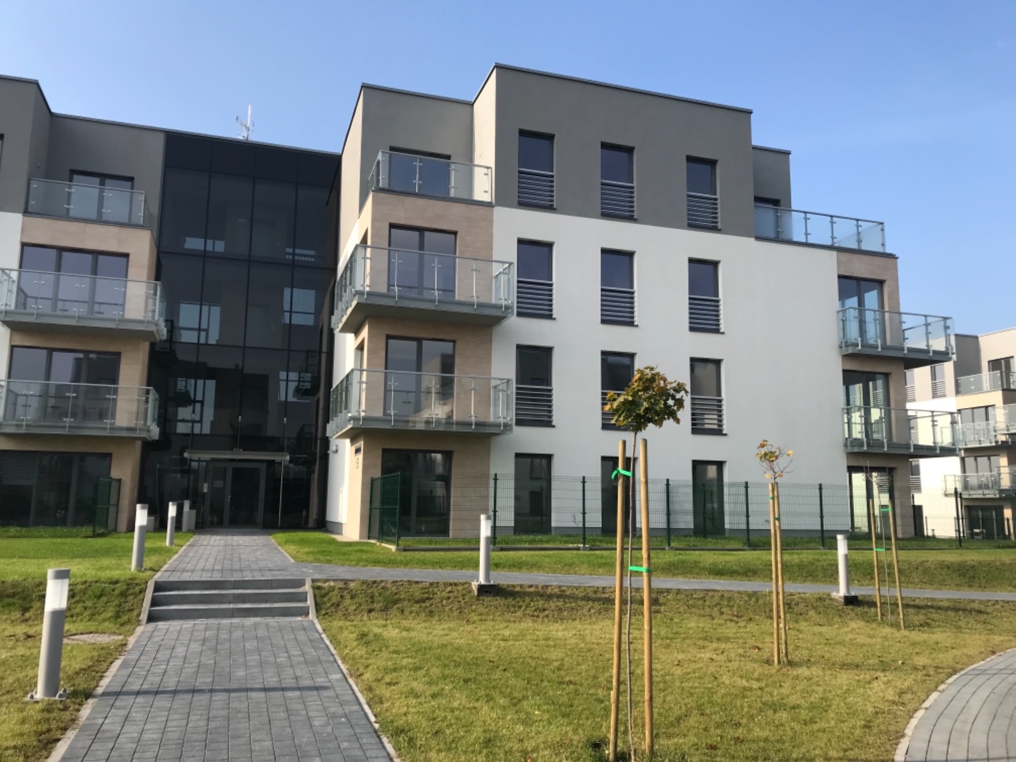 Nowoczesny budynek wielorodzinny z balkonami, elewacją w odcieniach szarości i bieli, oraz szklaną fasadą przy wejściu, otoczony trawnikiem z młodymi drzewkami.
