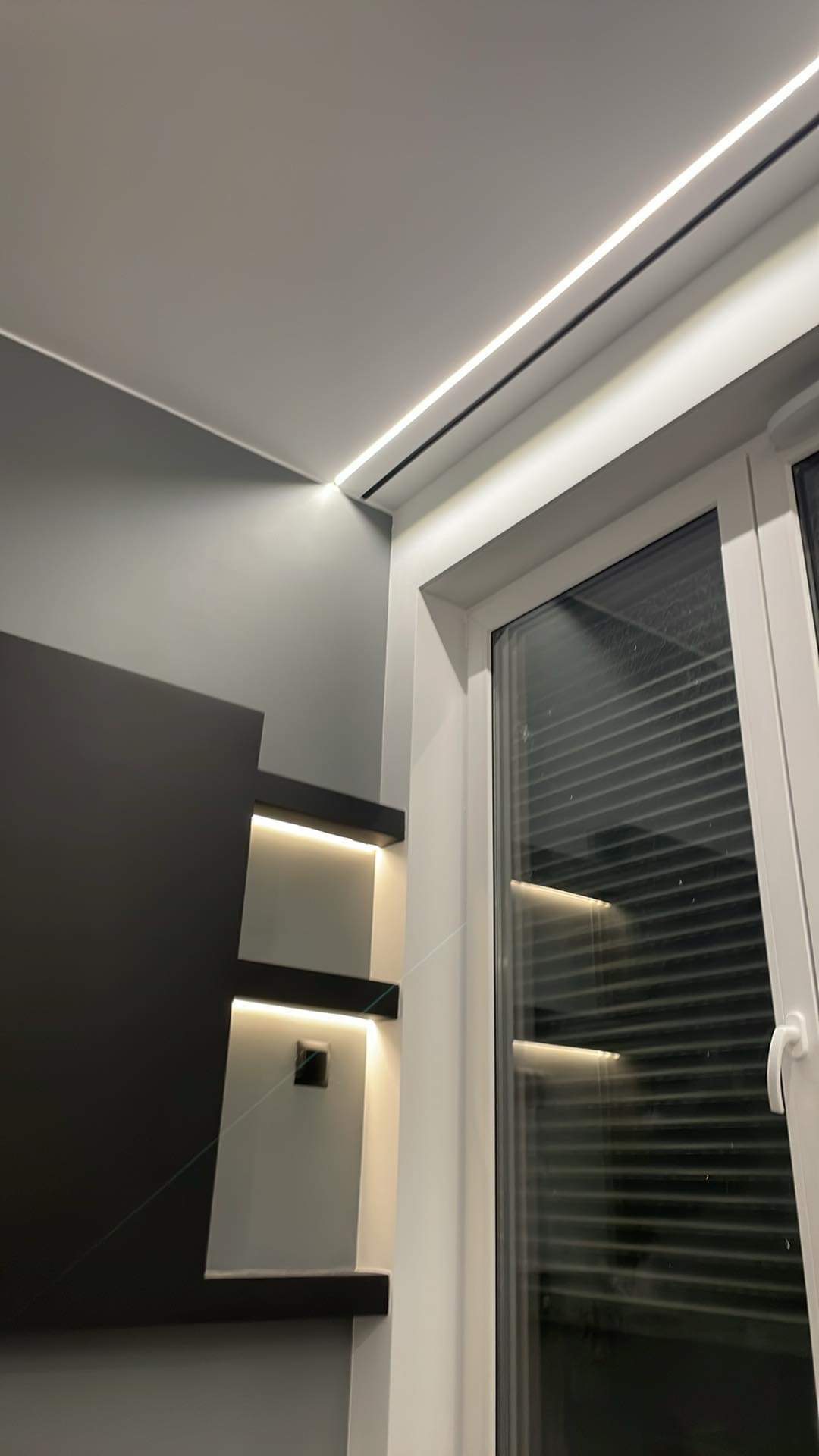 Nowoczesne oświetlenie LED wzdłuż sufitu i półek ściennych, podkreślające minimalistyczny design wnętrza z oknem z żaluzjami.