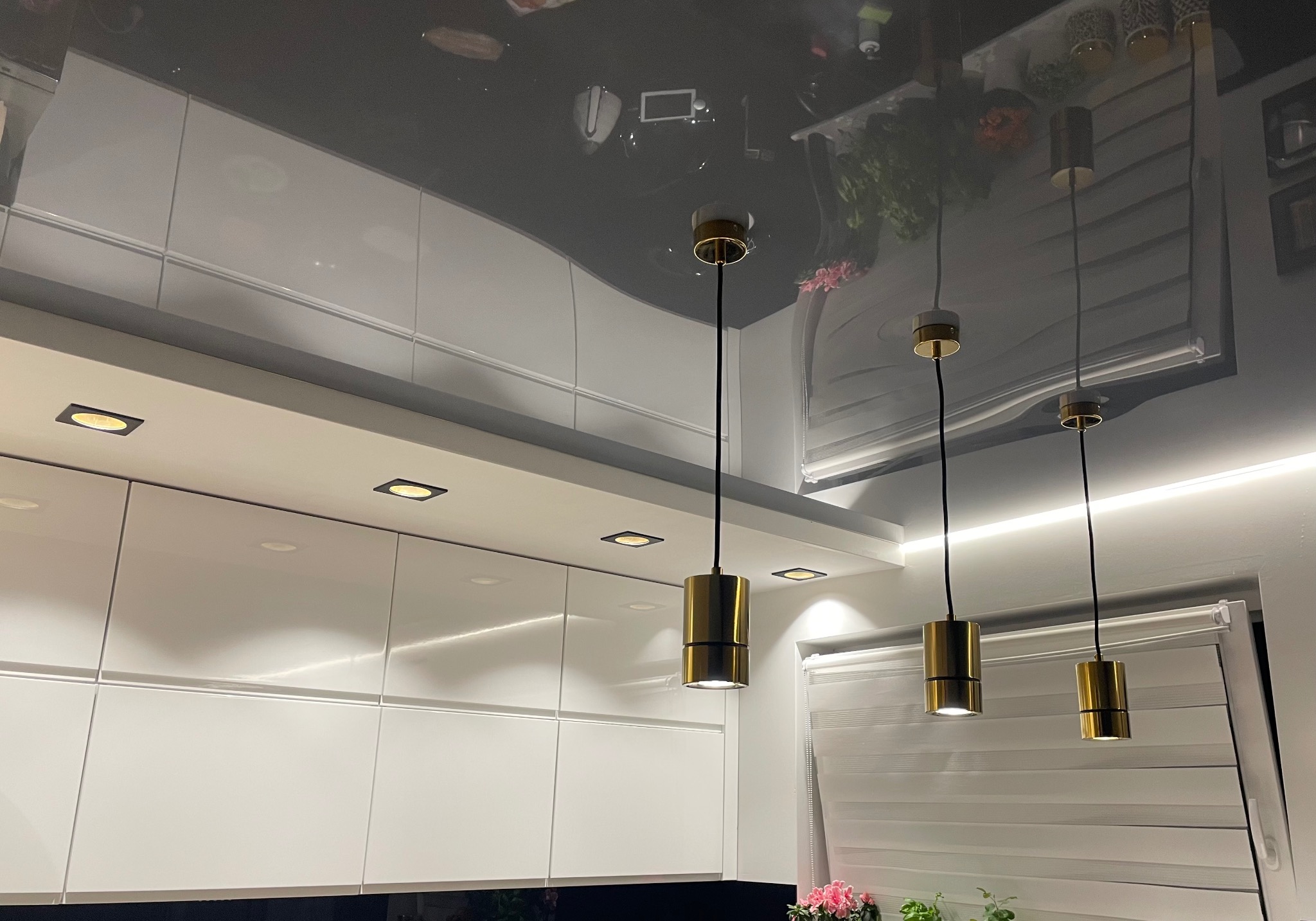 Nowoczesna kuchnia we Wrocławiu z białymi, błyszczącymi szafkami i trzema złotymi lampami wiszącymi, oświetlającymi blat. Widoczny fragment sufitu z odblaskiem.