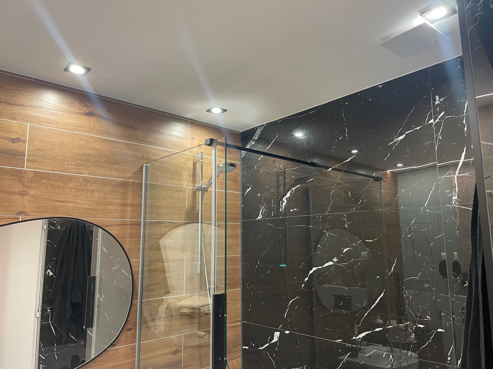 Nowoczesna kabina prysznicowa z czarnymi płytkami imitującymi marmur i drewnopodobnymi płytkami na ścianie, oświetlona punktowymi lampami sufitowymi, z widocznym okrągłym lustrem.