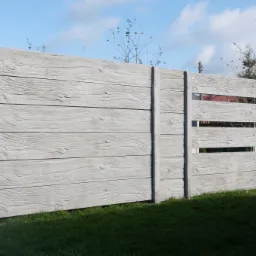 Szary betonowy płot z fakturą drewna, częściowo ażurowy, na tle zielonego trawnika i budynków.