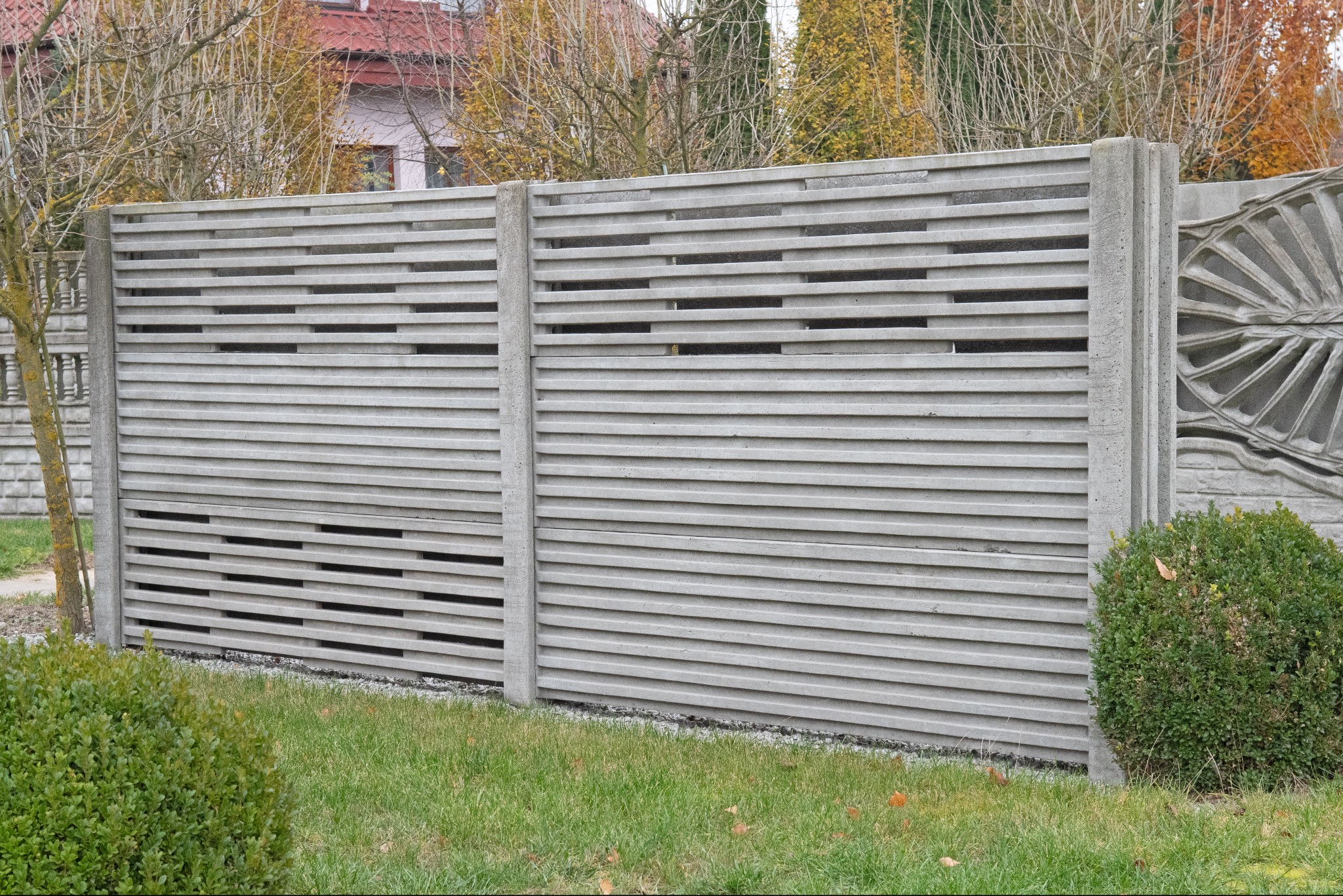 Fragment ogrodzenia z prefabrykatów betonowych, z poziomymi szczelinami, na tle trawnika i drzew z jesiennymi liśćmi.