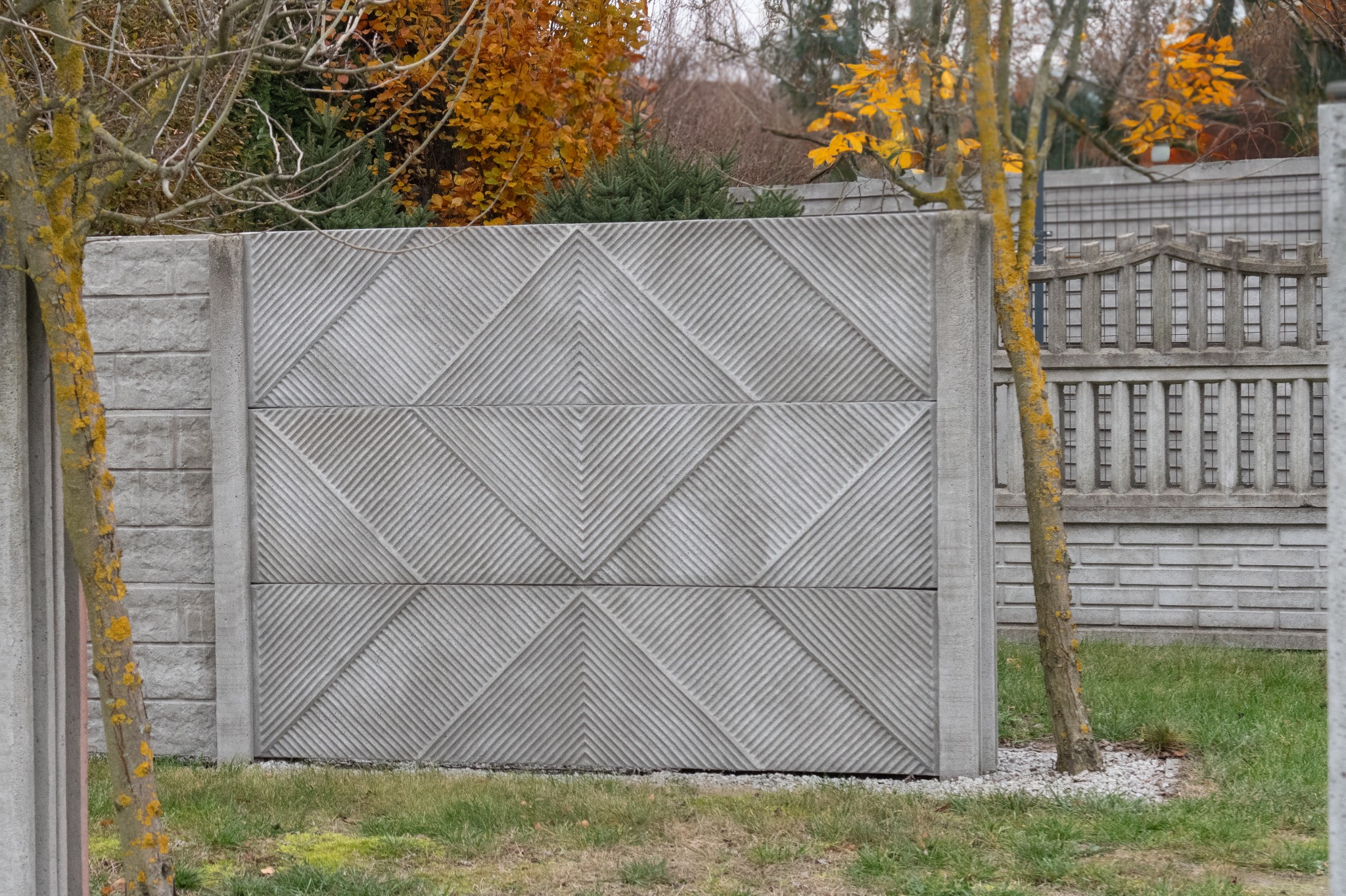 Szary, dwupanelowy płot betonowy z geometrycznym wzorem w romby, widoczny zza pnia drzewa pokrytego żółtym mchem, z fragmentem innego ogrodzenia w tle i jesiennymi drzewami.