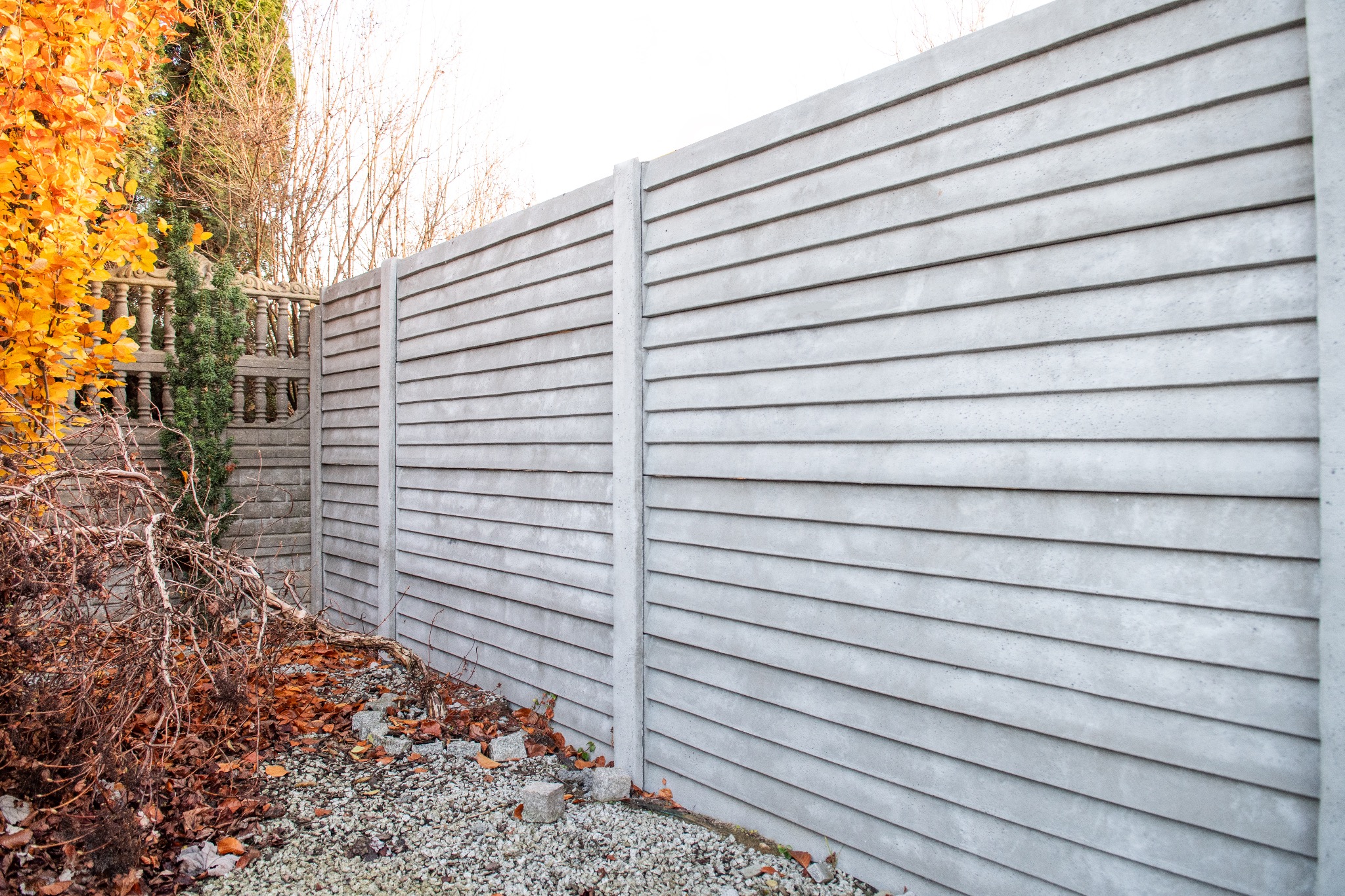 Szary, prefabrykowany płot z poziomych paneli betonowych, widoczny z bliska na tle jesiennego ogrodu z opadłymi liśćmi i fragmentem kamiennego murku.