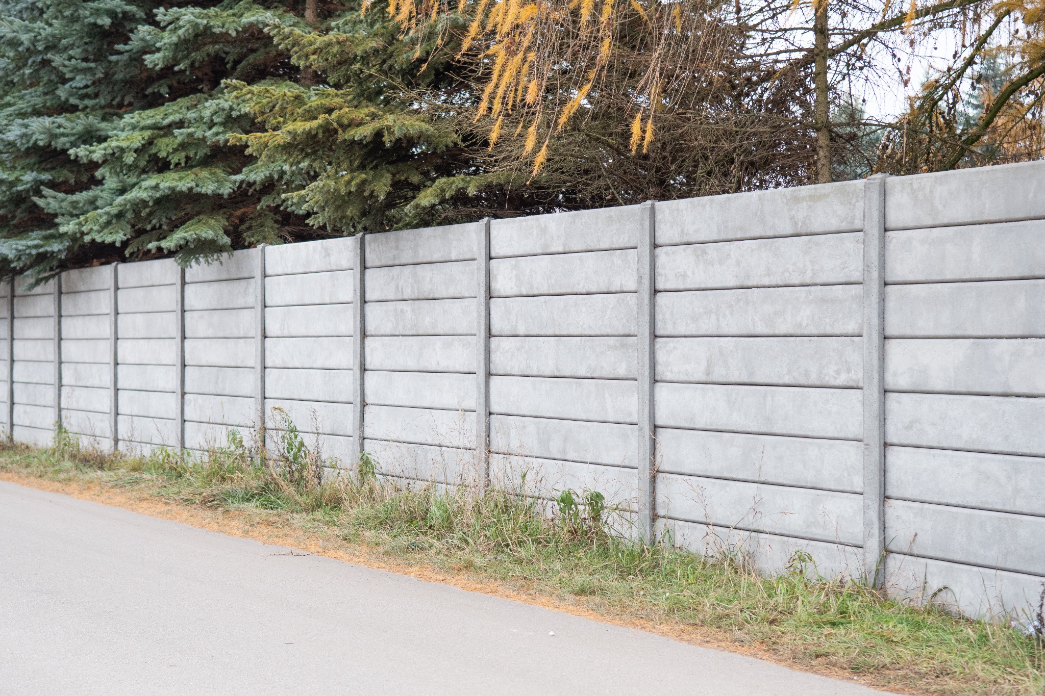 Długi, szary płot z prefabrykowanych elementów betonowych, widoczny z poziomu ulicy, z iglakami i drzewami w tle.