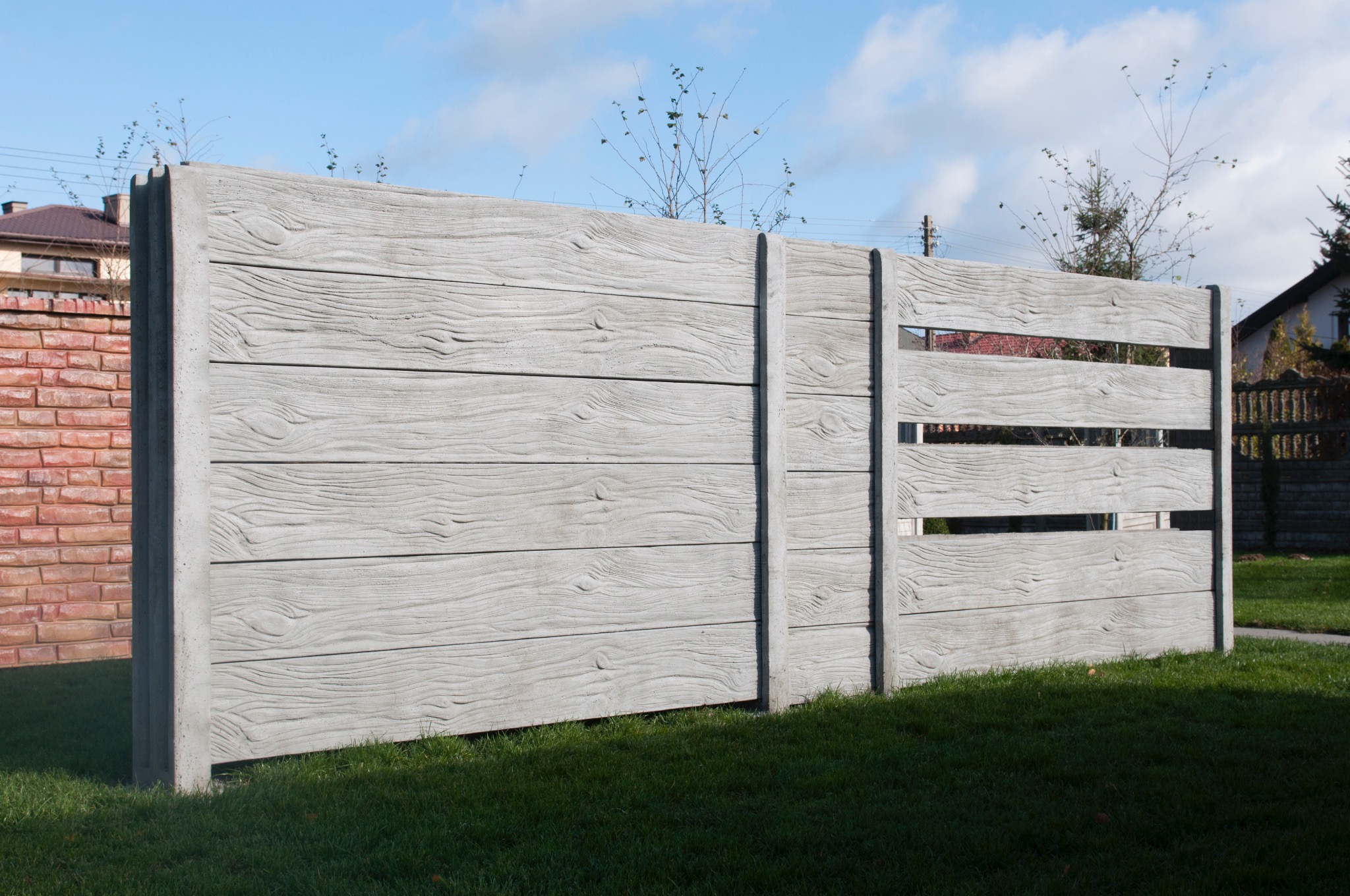 Szary betonowy płot z fakturą drewna, częściowo ażurowy, na tle zielonego trawnika i budynków.