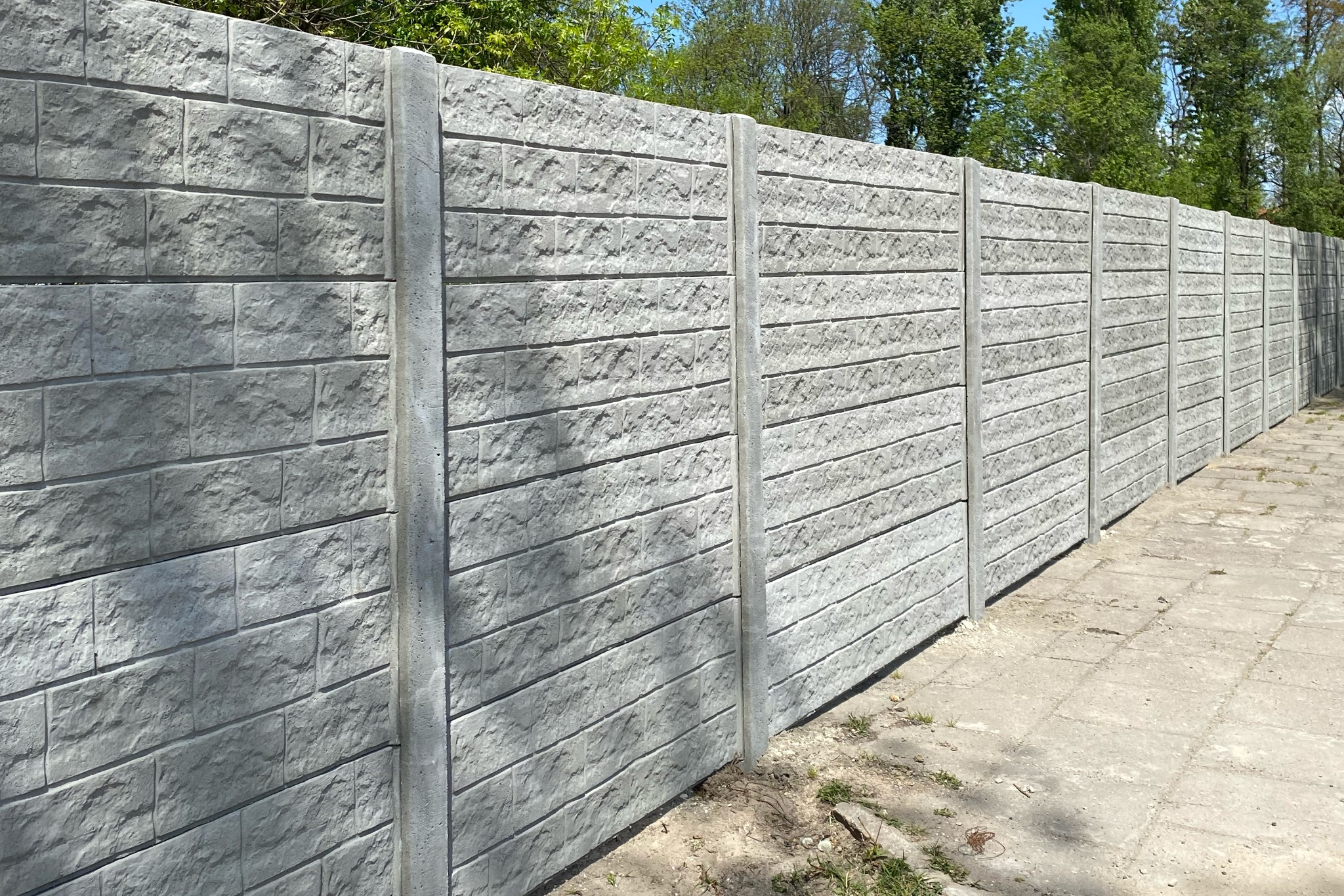 Długi, szary, prefabrykowany mur z paneli betonowych imitujących cegłę, oddzielonych pionowymi słupkami, ciągnący się wzdłuż chodnika.