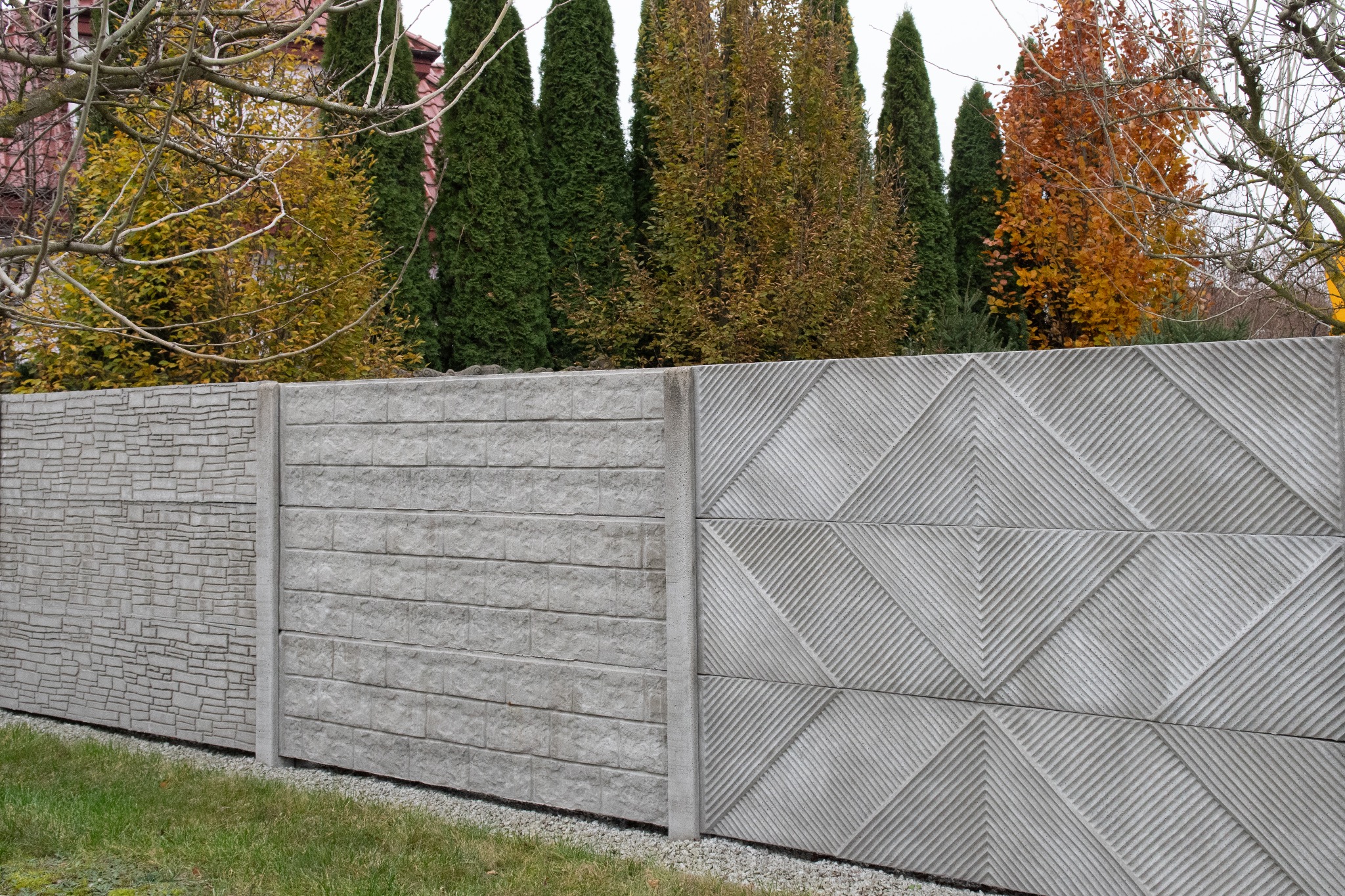 Fragment ogrodzenia z prefabrykatów betonowych o różnej fakturze, widoczne trzy przęsła: imitacja kamienia łupanego, cegły i wzór geometryczny w romby, w tle drzewa iglaste i liściaste.